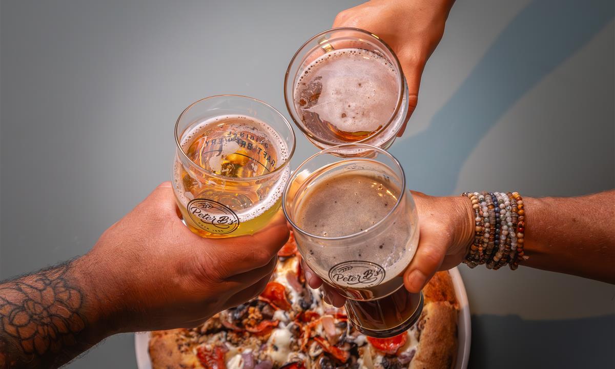 Three people clinking beers over a pizza