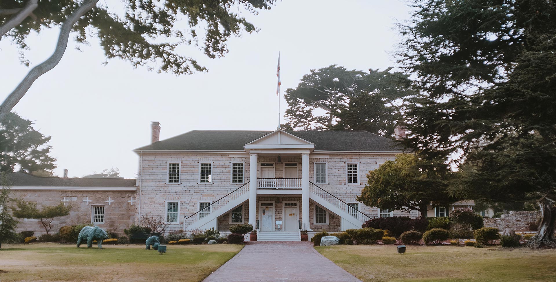 A two-story white brick building with a central staircase and columns, surrounded by lawn and trees.