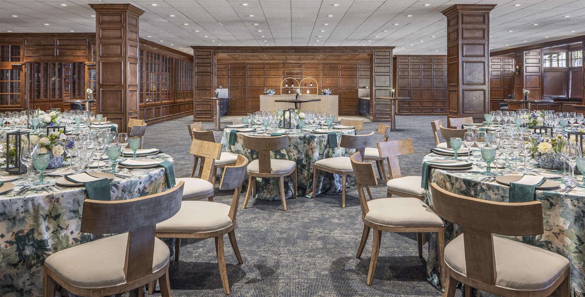A formal dining room with round tables set for a meal, featuring floral arrangements, glassware, and linens.