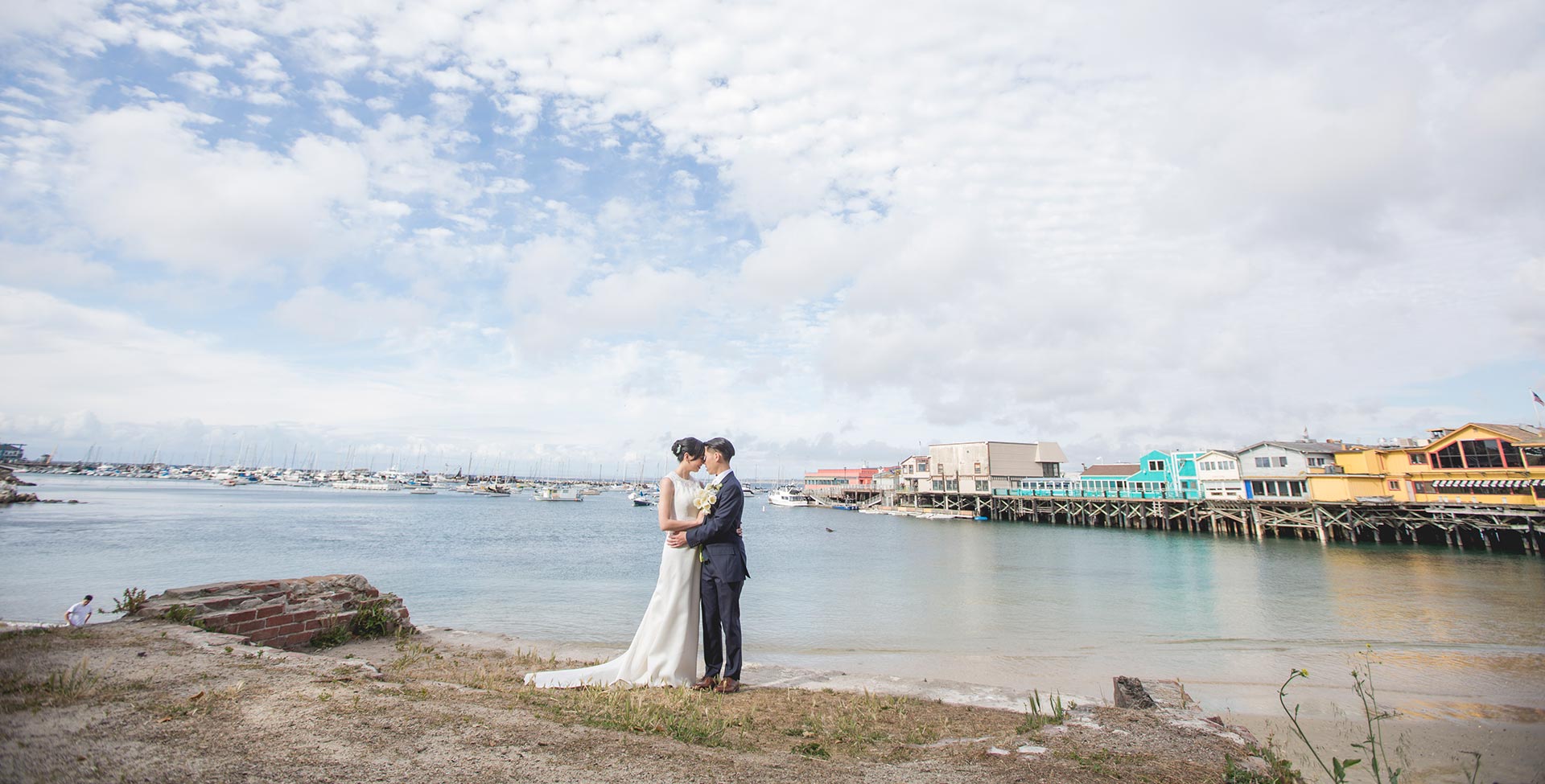 A couple in formal attire embracing on a beach with a body of water and a pier in the background.